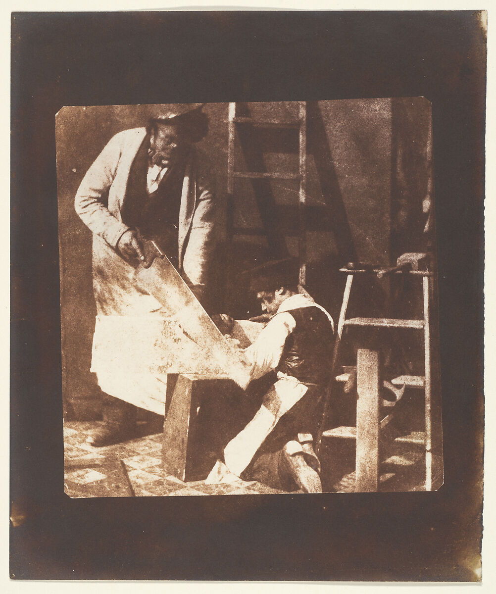 A carpenter guides a kneeling apprentice as they work together at a bench.
